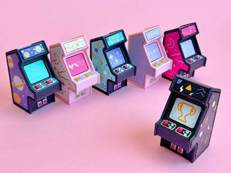 Six mini arcade cabinet clickers with printed side art on a pink background